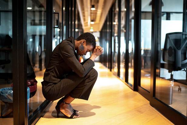 A Black woman crouches in a hallway with her head in her hands. She may be experiencing work stress.